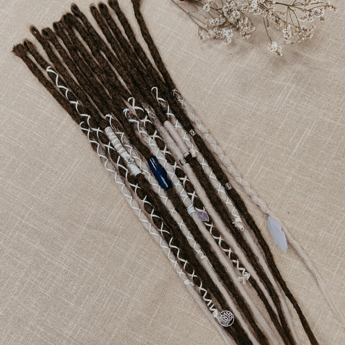 A set of long dreadlocks styled with white braided yarn, small beads, and decorative stones, laid flat on a textured beige fabric background.