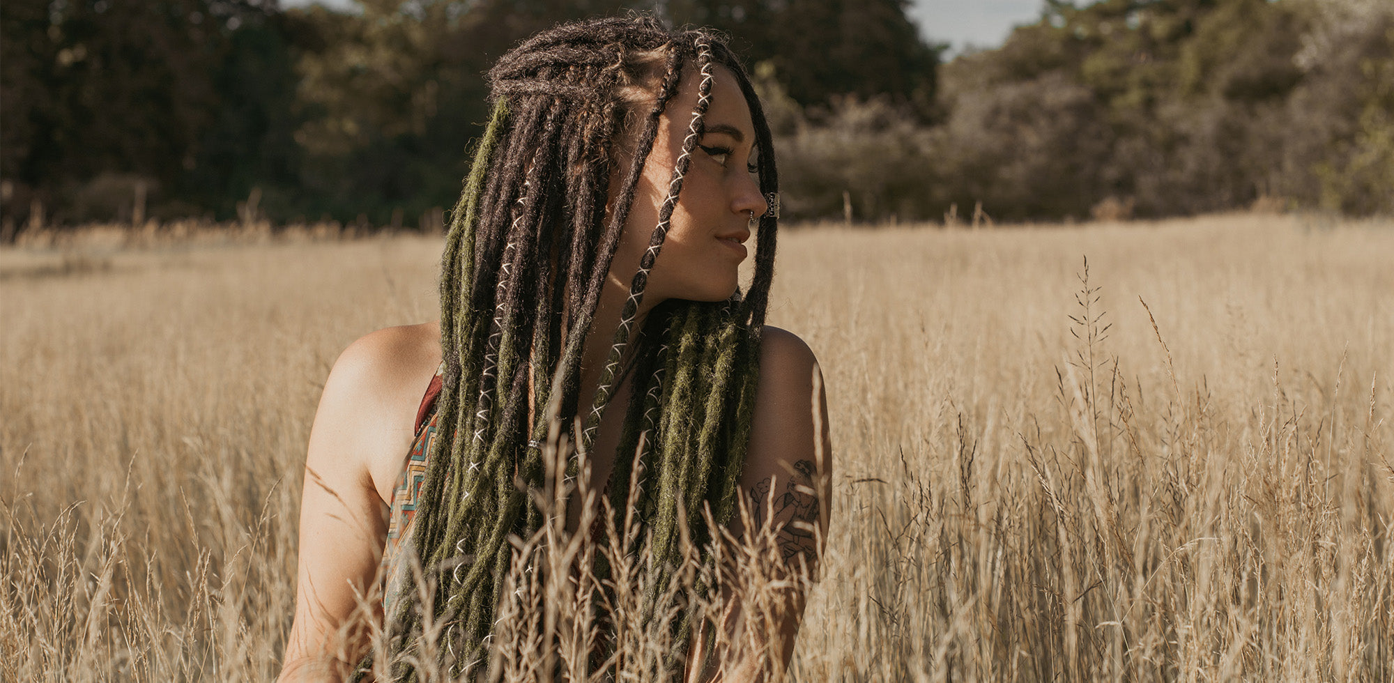Romy wearing her Dark green dreads (mossy) sitting in a field and looking to the right