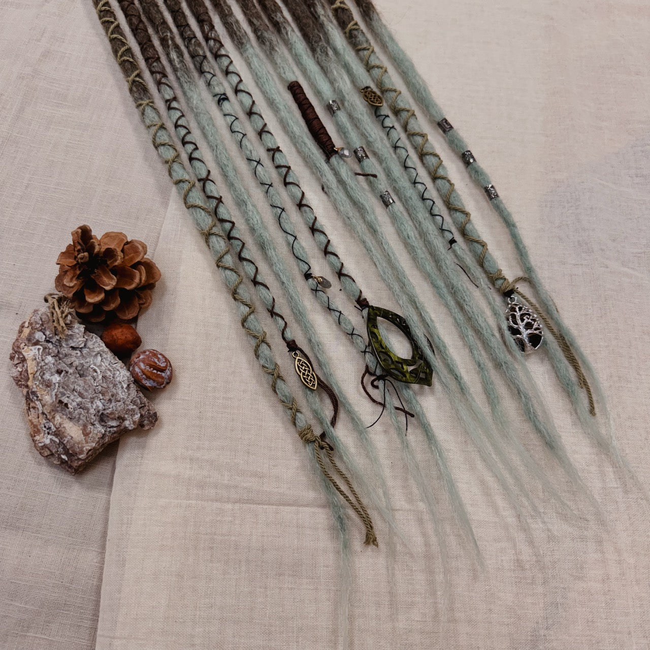 A set of handmade dreadlock extensions, laid flat on a textured beige fabric background, featuring a dark brown to light blue-green ombré color scheme with decorative braided and wavy details.