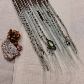 A set of handmade dreadlock extensions, laid flat on a textured beige fabric background, featuring a dark brown to light blue-green ombré color scheme with decorative braided and wavy details.
