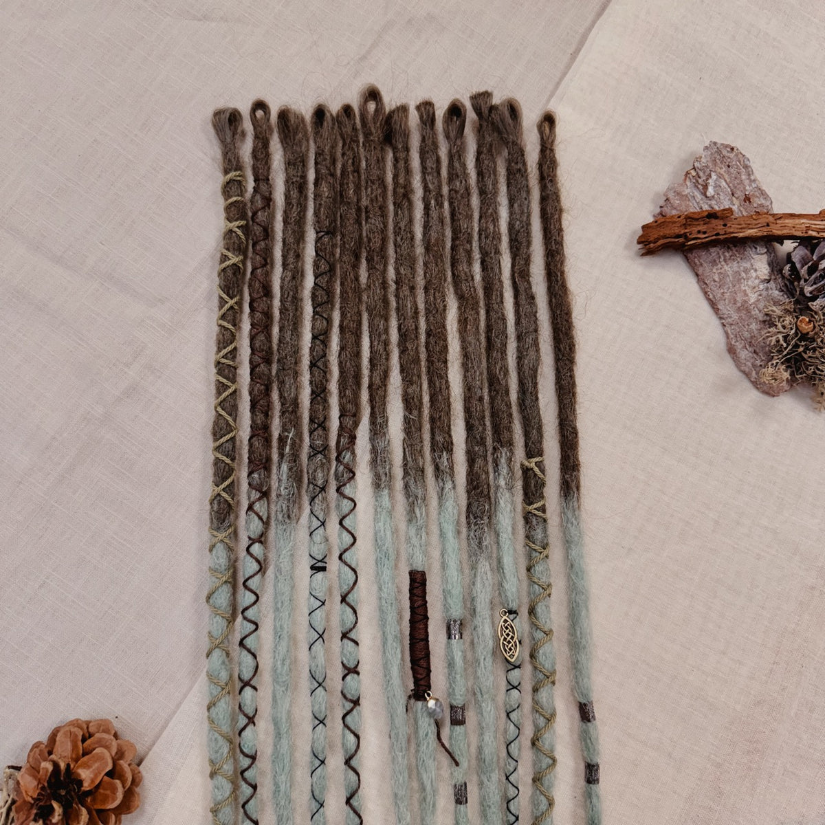 A set of handmade dreadlock extensions, laid flat on a textured beige fabric background, featuring a dark brown to light blue-green ombré color scheme with decorative braided and wavy details.