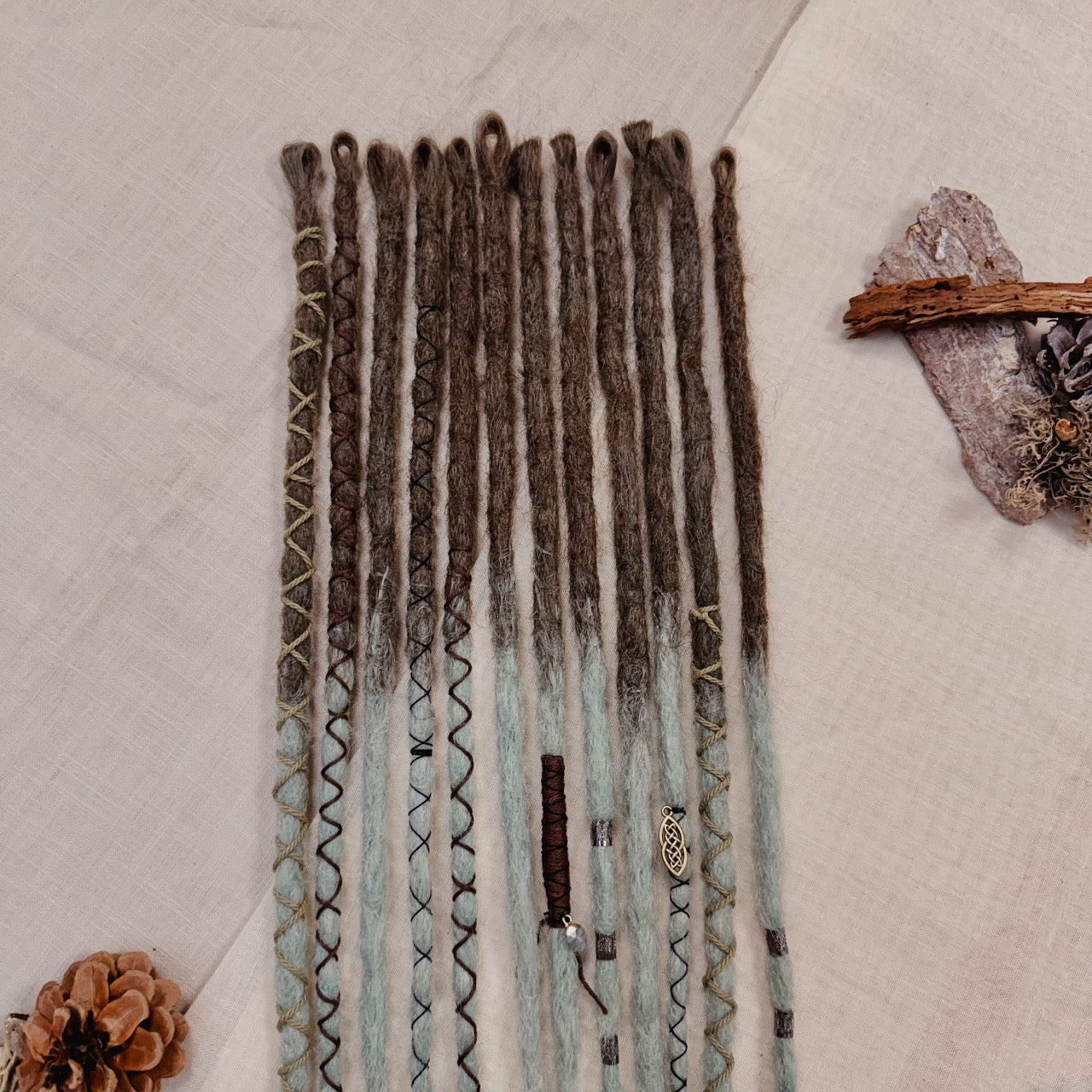 A set of handmade dreadlock extensions, laid flat on a textured beige fabric background, featuring a dark brown to light blue-green ombré color scheme with decorative braided and wavy details.