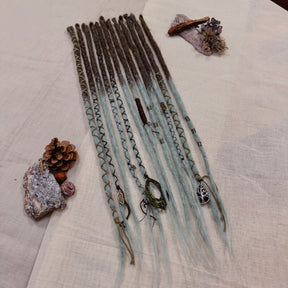 A set of handmade dreadlock extensions, laid flat on a textured beige fabric background, featuring a dark brown to light blue-green ombré color scheme with decorative braided and wavy details.