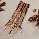 A set of long, multi-colored synthetic dreadlock extensions decorated with yarn and beads laid out on a off-white fabric background, with dried botanicals and small stones around it. The photo shows the whole set