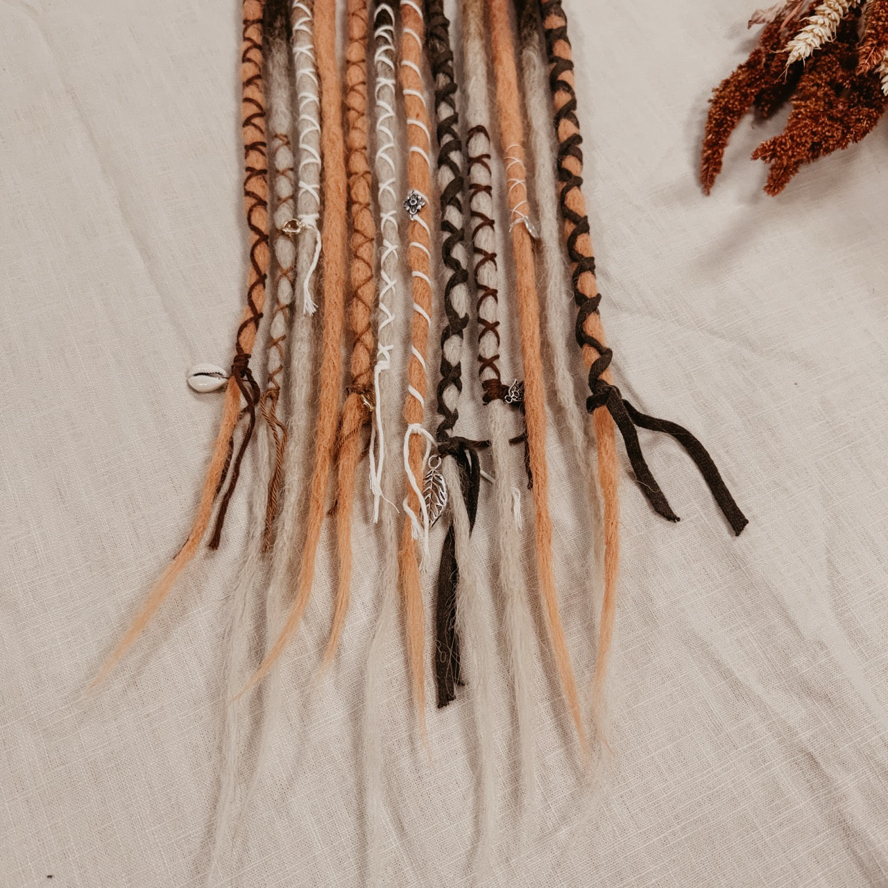 A set of long, multi-colored synthetic dreadlock extensions decorated with yarn and beads laid out on a off-white fabric background, with dried botanicals and small stones around it. The photo shows the bottom part of the set