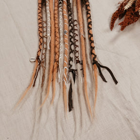 A set of long, multi-colored synthetic dreadlock extensions decorated with yarn and beads laid out on a off-white fabric background, with dried botanicals and small stones around it. The photo shows the bottom part of the set