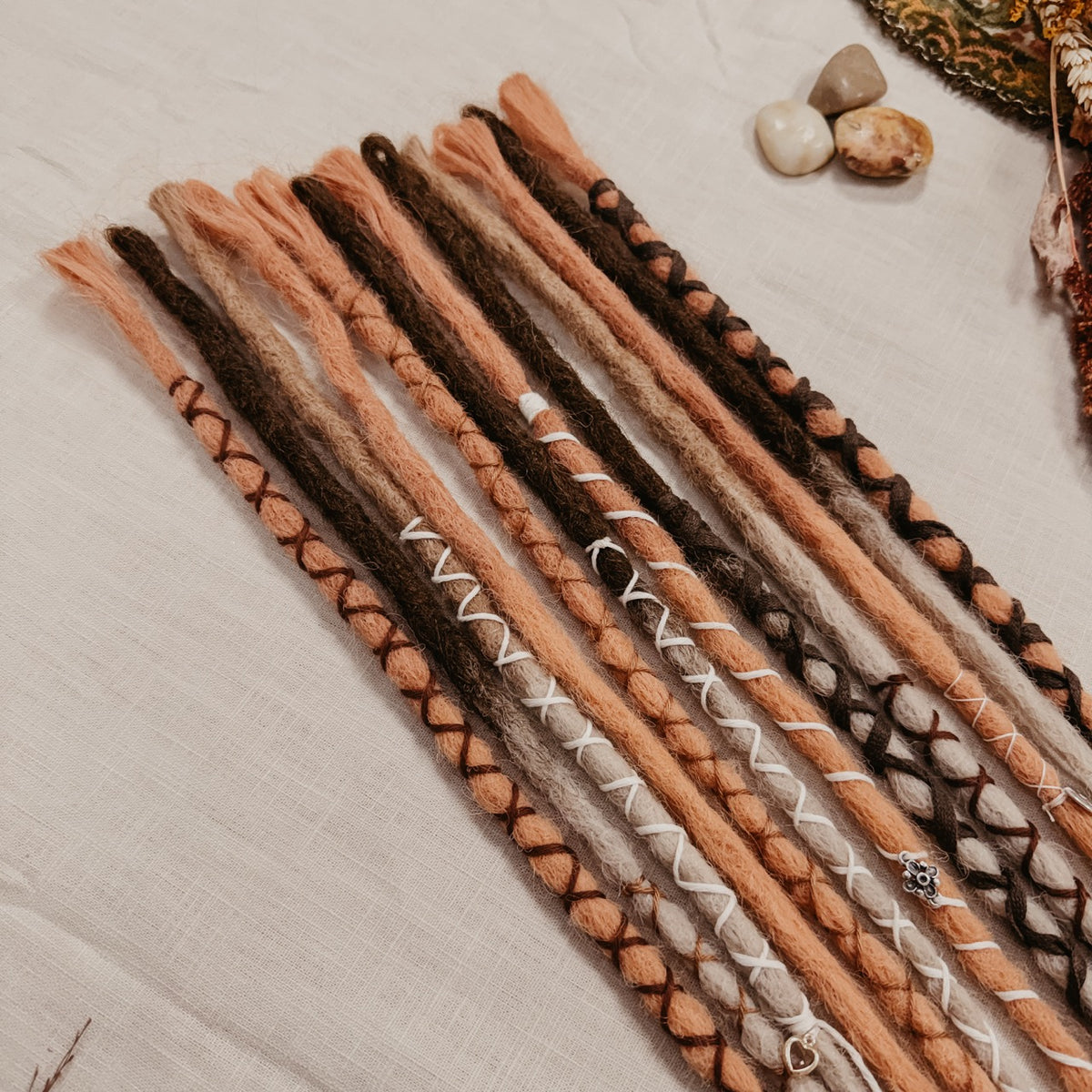 A set of long, multi-colored synthetic dreadlock extensions decorated with yarn and beads laid out on a off-white fabric background, with dried botanicals and small stones around it. The photo shows the top part of the set