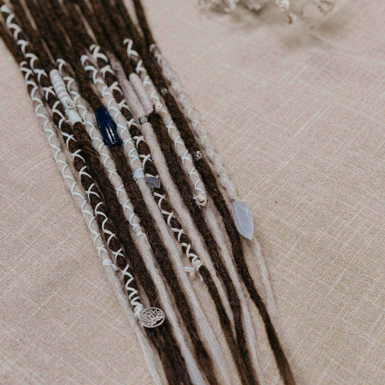 A close-up of decorated dreadlock set with dark brown and ombre white synthetic dreadlocks, adorned with white blue and silver  beads, laid on a beige linen background.