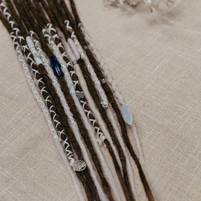 A close-up of decorated dreadlock set with dark brown and ombre white synthetic dreadlocks, adorned with white blue and silver  beads, laid on a beige linen background.