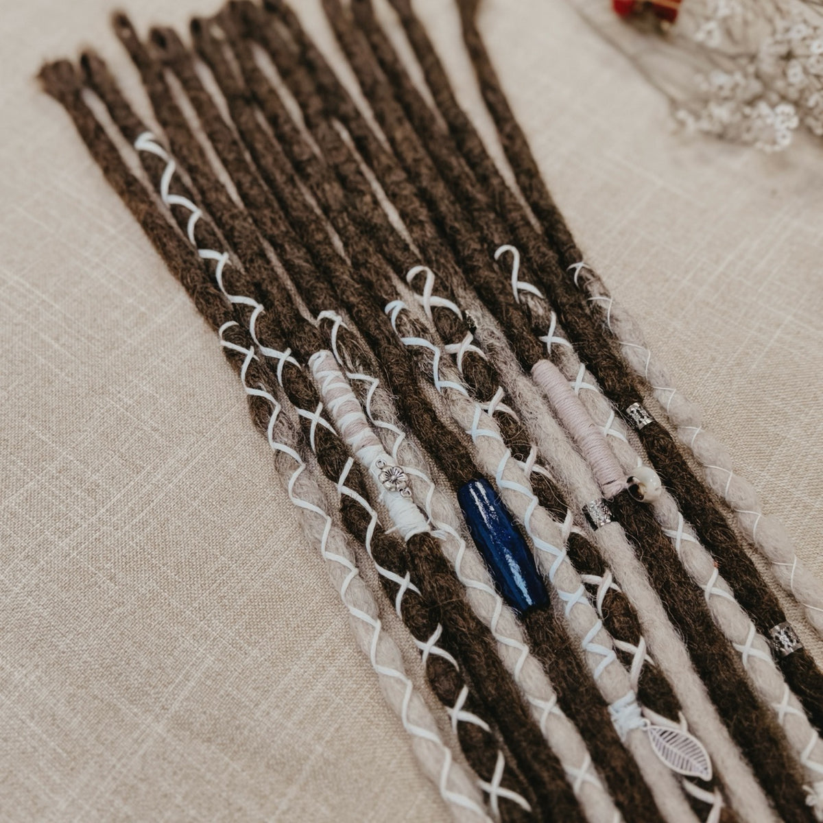 A close-up of decorated dreadlock set with dark brown and ombre white synthetic dreadlocks, adorned with white blue and silver  beads, laid on a beige linen background.