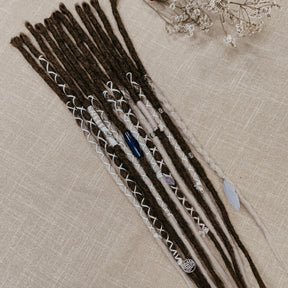 A set of long dreadlocks styled with white braided yarn, small beads, and decorative stones, laid flat on a textured beige fabric background.