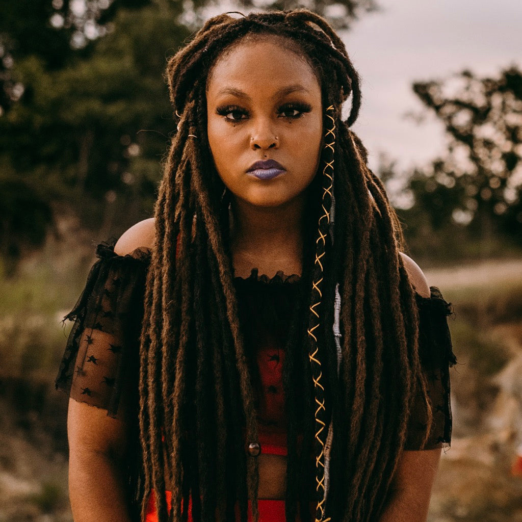 Woman with long dreadlock extensions, standing outdoors with trees in the background