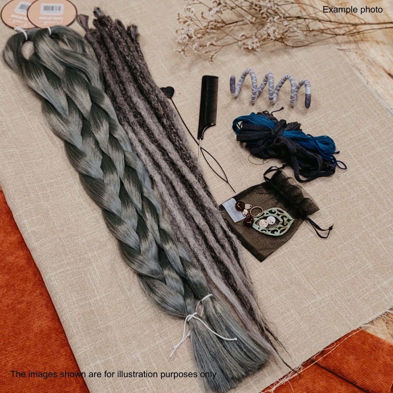 A styled flat-lay of hair extension materials on a beige fabric surface, including a long braided synthetic dreadlock sample, several loose synthetic dreadlocks, a black comb, two coiled bundles of blue and black yarn, a small pouch, and a decorative tag. Text in the corners says 'Example photo' and 'The images shown are for illustration purposes only.